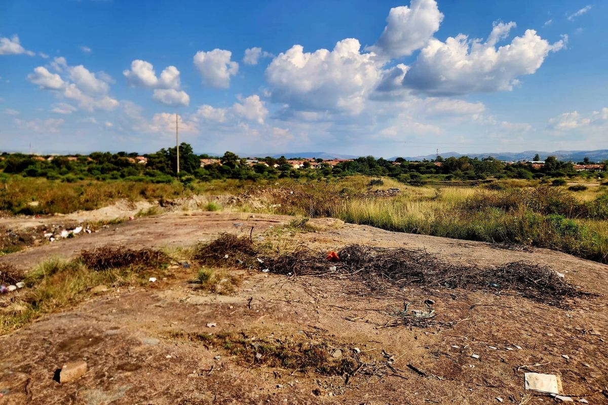 A view of an undeveloped residential plot featuring natural terrain, scrub vegetation, and distant suburban housing under a clear blue sky.