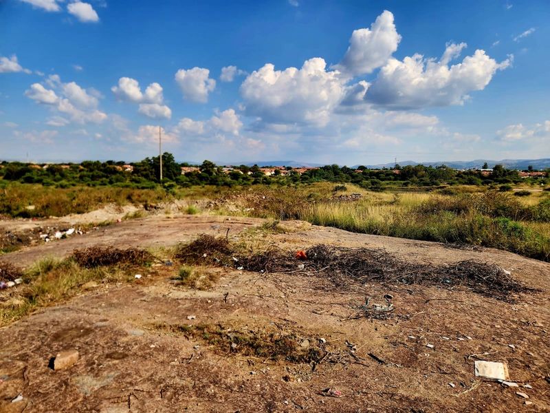 A view of an undeveloped residential plot featuring natural terrain, scrub vegetation, and distant suburban housing under a clear blue sky.