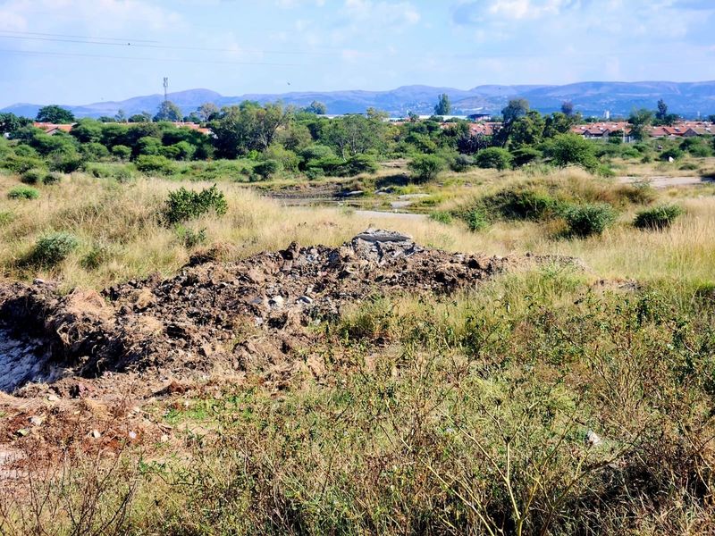 A view of a vacant residential plot featuring natural vegetation and a pile of debris, with distant mountain and suburban views.