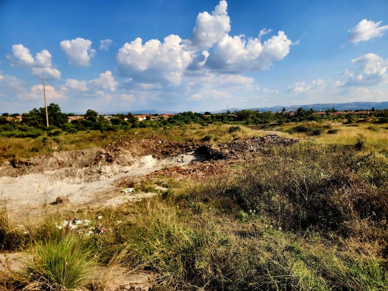 An undeveloped residential land plot featuring natural vegetation and uneven terrain under a clear blue sky.