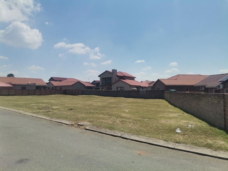 A vacant residential plot of land featuring a level grass surface, enclosed by a perimeter brick wall, situated in a developed suburban neighborhood.