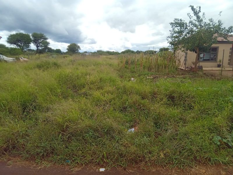 A view of an undeveloped residential plot featuring overgrown grass and natural vegetation, adjacent to an existing structure.