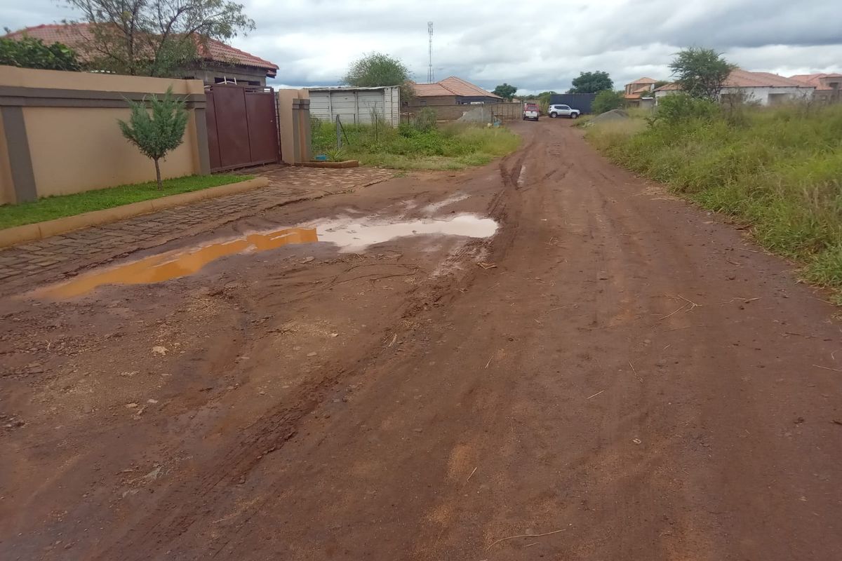 A view of an unpaved, muddy access road leading to a residential property with a boundary wall and gated entrance.