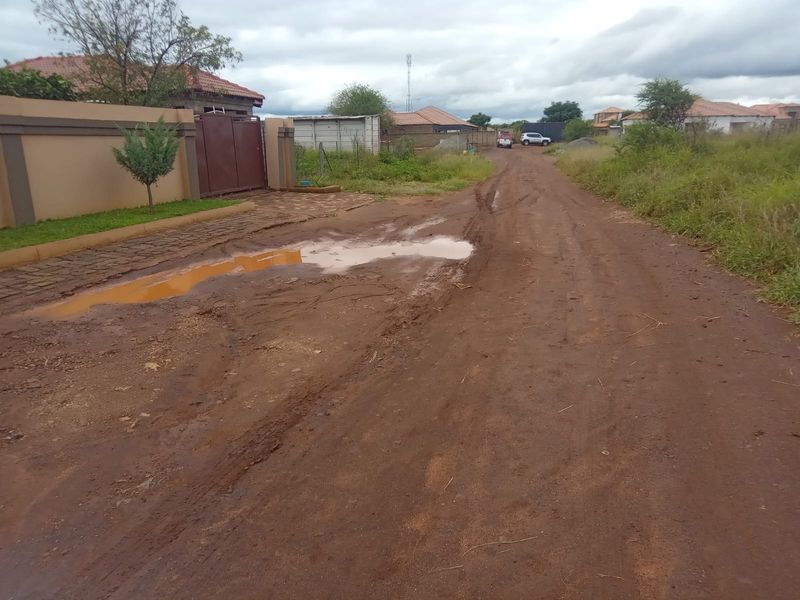 A view of an unpaved, muddy access road leading to a residential property with a boundary wall and gated entrance.