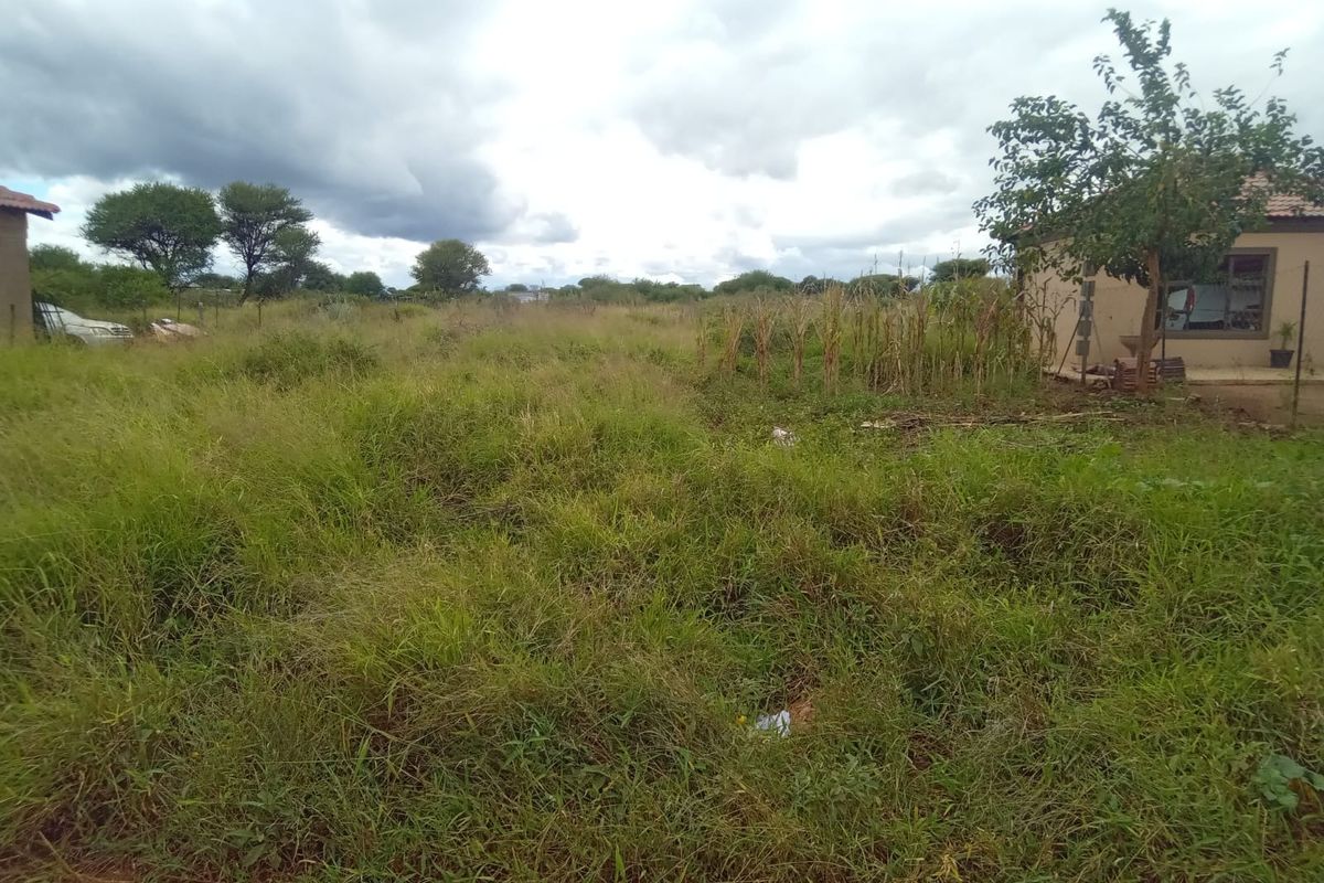 A view of an undeveloped residential plot featuring overgrown grass and natural vegetation with adjacent structures visible in the background.