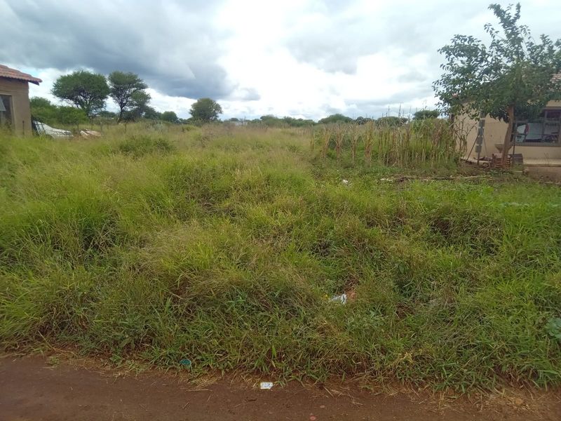 A view of an undeveloped residential plot featuring overgrown grass and natural vegetation, situated between existing neighboring structures.
