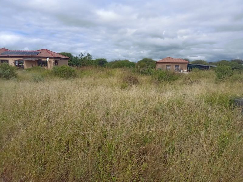 A view of a large residential plot featuring overgrown grass and two existing residential structures with tiled roofs and solar panels.