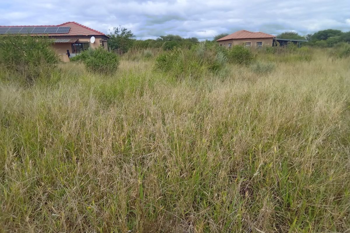 A view of a residential plot featuring tall, overgrown grass with existing structures visible in the background under a cloudy sky.
