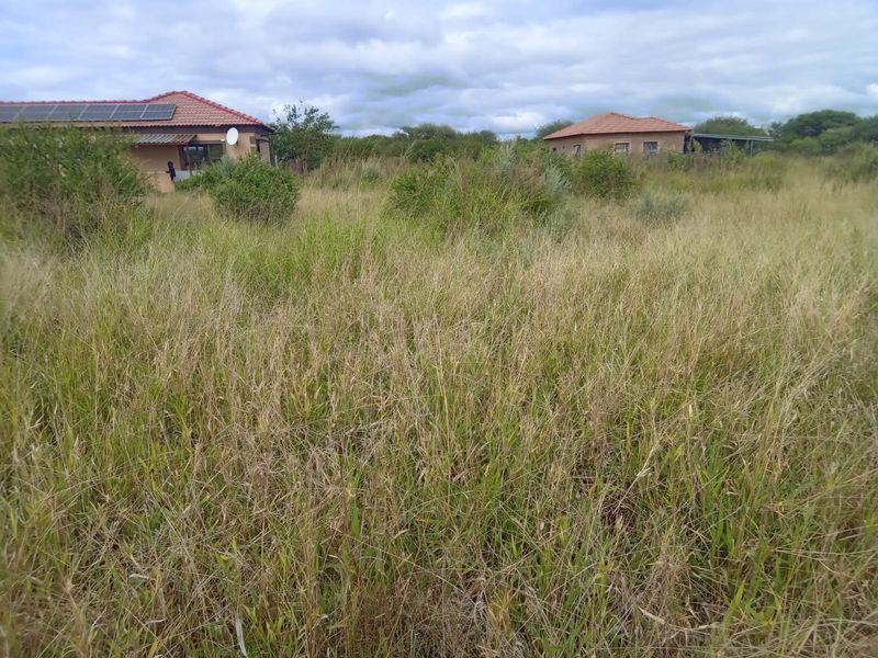 A view of a residential plot featuring tall, overgrown grass with existing structures visible in the background under a cloudy sky.