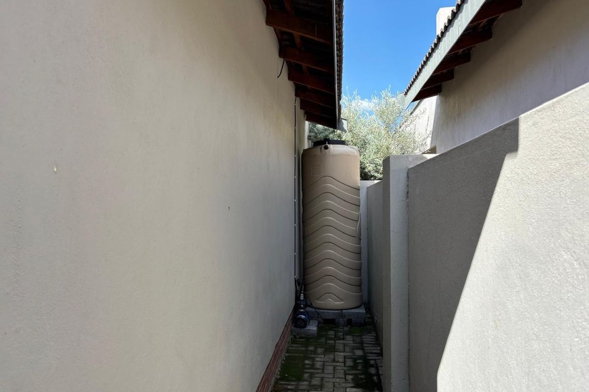 A narrow side passage featuring a water storage tank and pump, with paved flooring and boundary walls between two building structures.