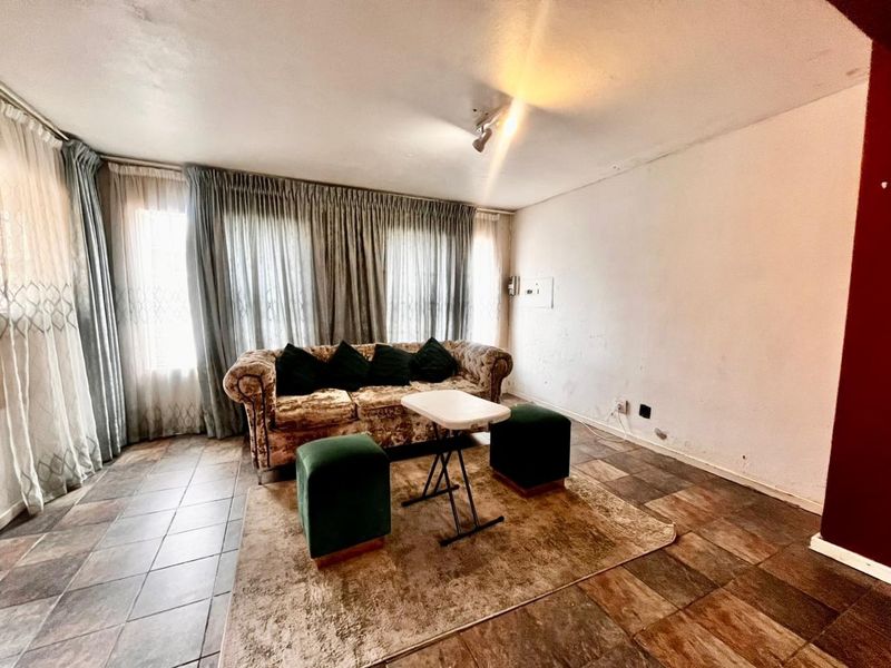 A living room area featuring tiled flooring and large windows with curtain tracks, requiring cosmetic updates to the walls and flooring.