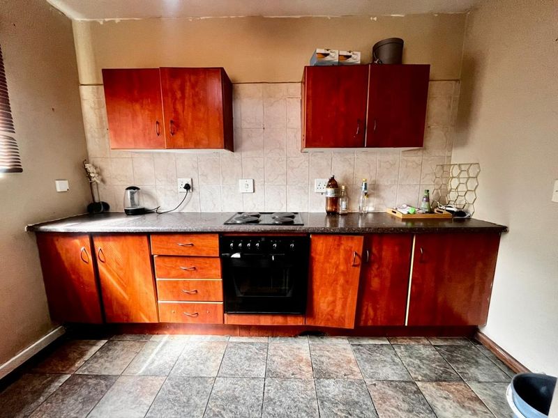 A functional kitchen featuring wooden cabinetry, a built-in oven and hob, tiled splashback, and durable tiled flooring, offering potential for modern cosmetic updates.