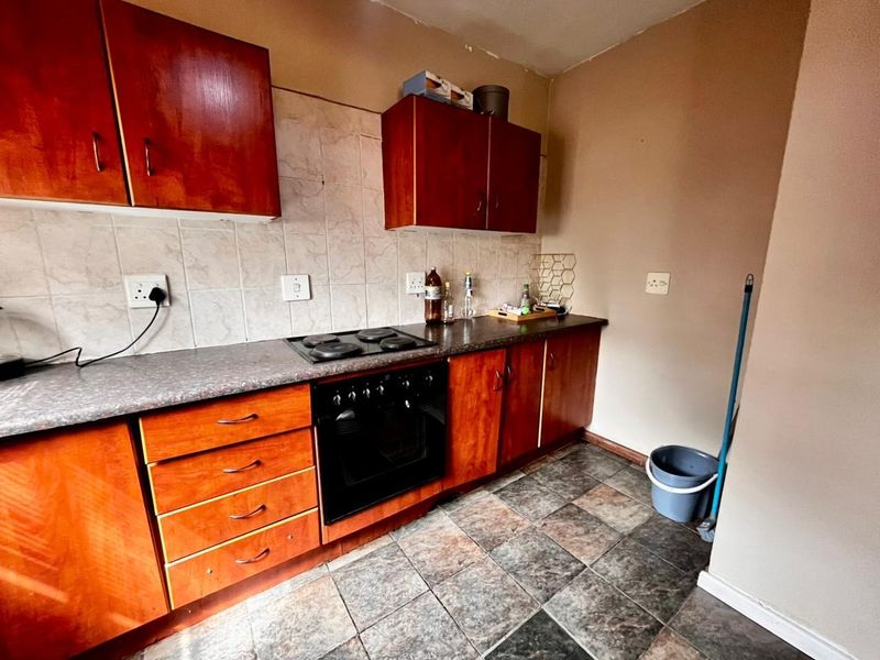 A functional kitchen featuring wooden cabinetry, a built-in oven with electric hob, tiled backsplash, and durable tiled flooring, offering potential for modern updates.