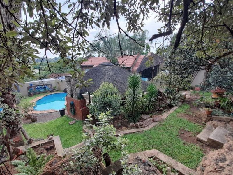 Outdoor pool area featuring a thatched lapa, brick fireplace, and tiered garden landscaping with stone retaining walls.