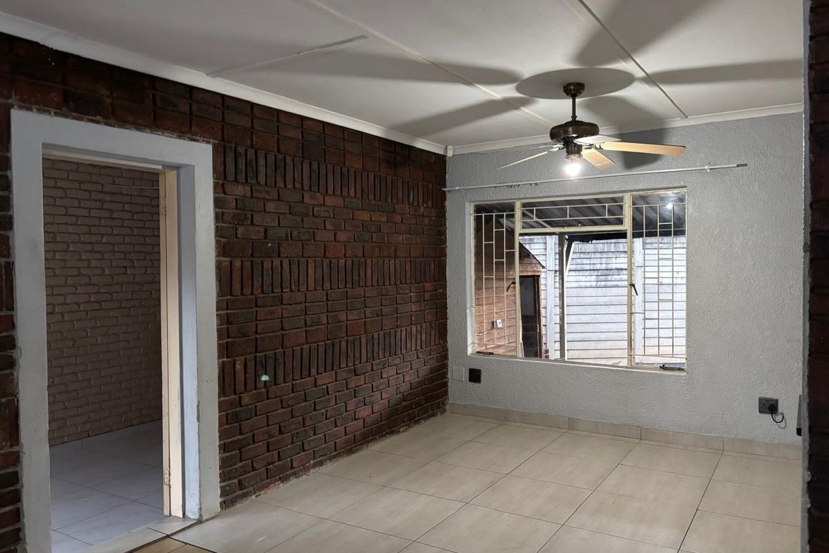 An empty living room featuring an exposed brick feature wall, tiled flooring, and a ceiling fan, offering a blank canvas for personalization.