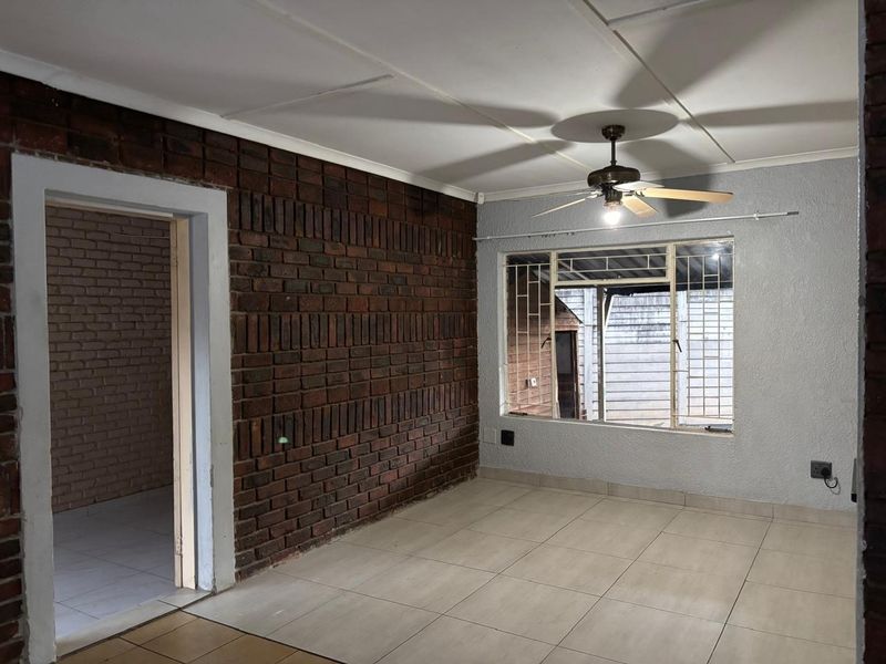 An empty living room featuring an exposed brick feature wall, tiled flooring, and a ceiling fan, offering a blank canvas for personalization.