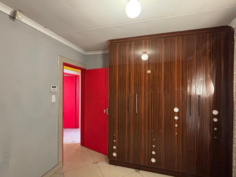 A bedroom featuring a large built-in wooden wardrobe, tiled flooring, and an alarm system keypad, with access to an adjacent hallway.