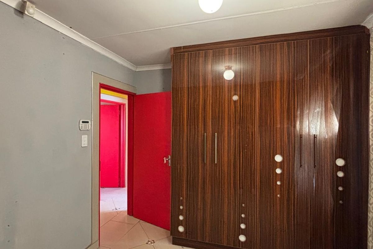 A bedroom featuring a large built-in wooden wardrobe, tiled flooring, and an alarm system keypad, with access to an adjacent hallway.