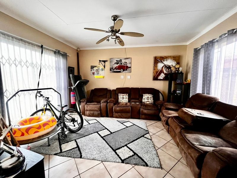 A living room featuring tiled flooring, a ceiling fan, and large windows with security gates, offering a functional space for relaxation.
