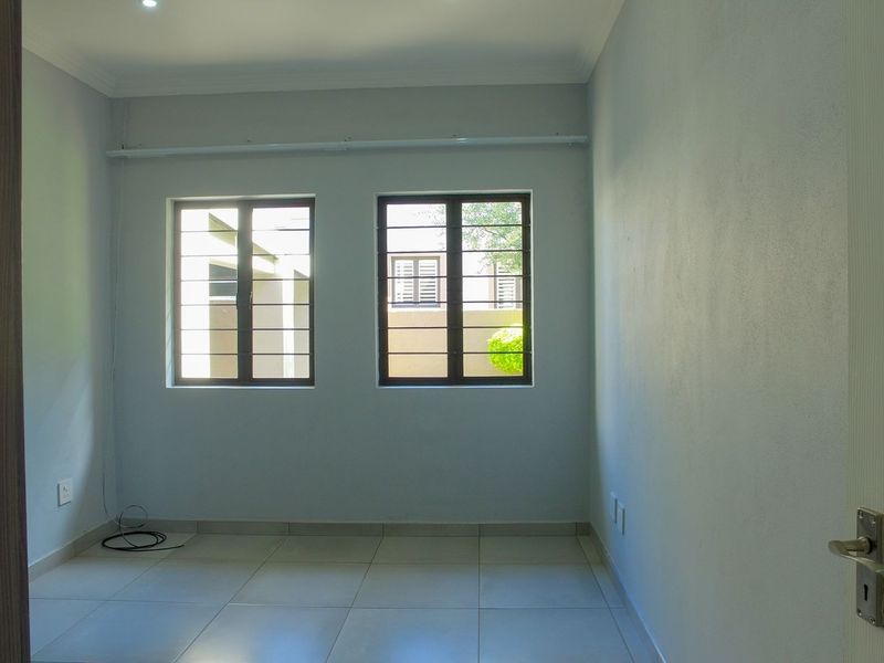 A clean, empty bedroom featuring neutral tiled flooring, built-in cupboards, and windows fitted with security bars.