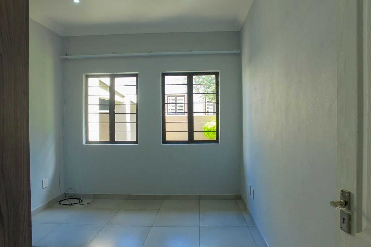 A clean, empty bedroom featuring neutral tiled flooring, built-in cupboards, and windows fitted with security bars.