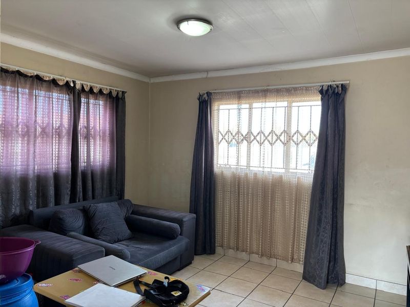 A living room featuring tiled flooring, security bars on windows, and standard ceiling lighting, requiring cosmetic updates to improve its overall appeal.