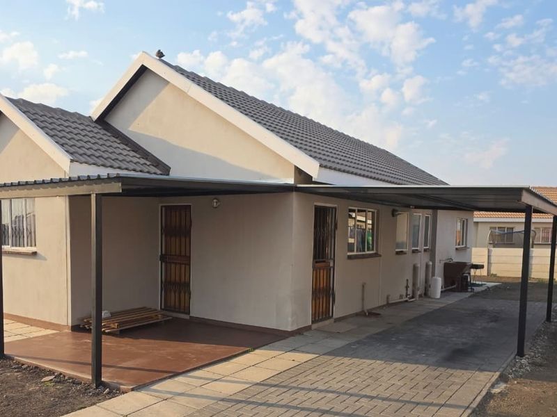 Rear view of the house featuring a covered patio, paved driveway, metal carport structure, and secure access points with security gates.