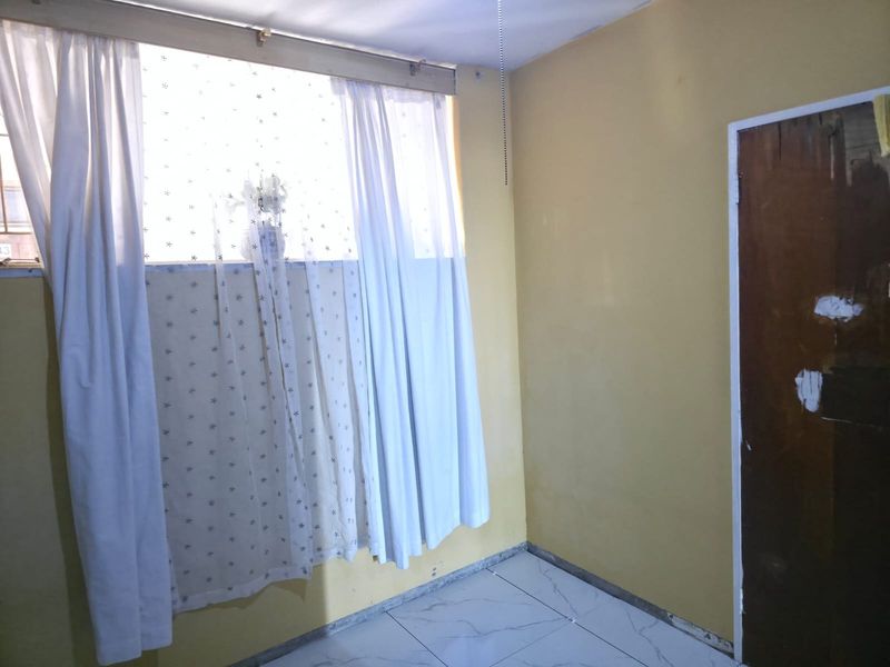 An empty bedroom featuring tiled flooring, a timber door, and a window. The room requires cosmetic updates including painting and trim installation.