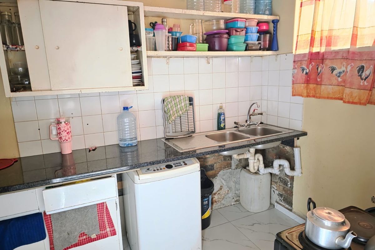 A functional kitchen area featuring a double bowl sink, tiled splashback, and basic cabinetry, currently showing signs of wear and exposed plumbing.