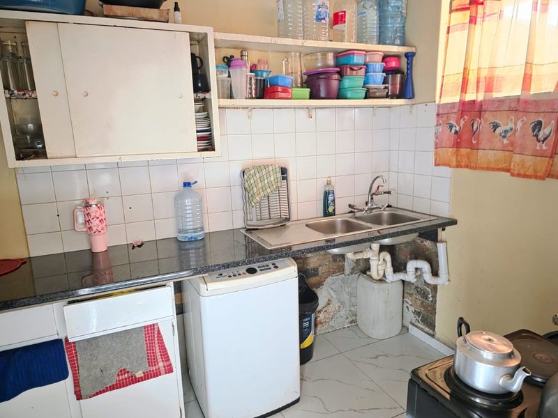 A functional kitchen area featuring a double bowl sink, tiled splashback, and basic cabinetry, currently showing signs of wear and exposed plumbing.