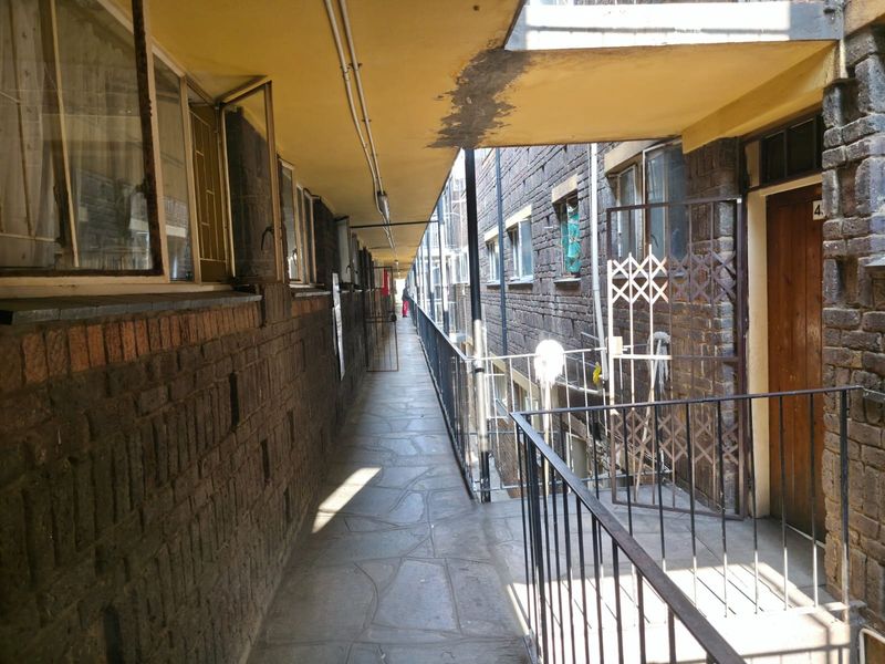 An exterior communal walkway featuring exposed brick walls, stone paving, and metal security gates, showing signs of wear and maintenance requirements.