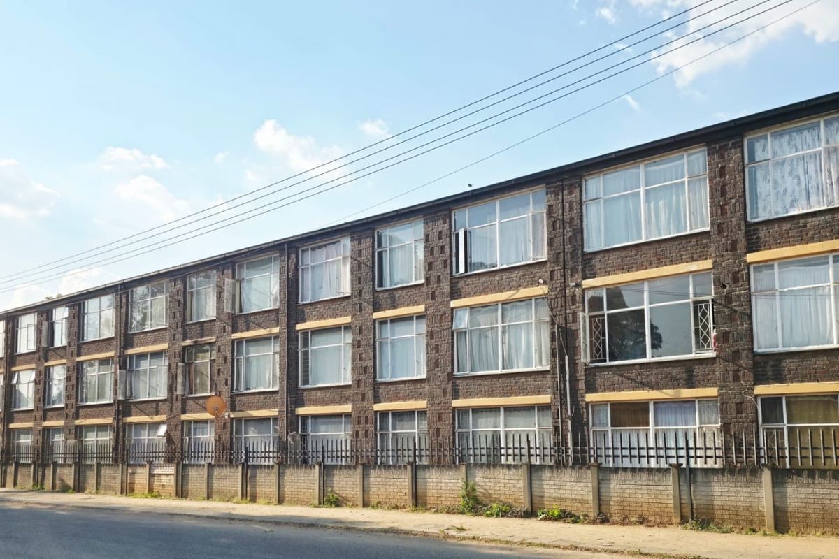 A multi-story residential apartment building featuring a classic brick facade, large metal-framed windows, and a secure perimeter boundary wall.