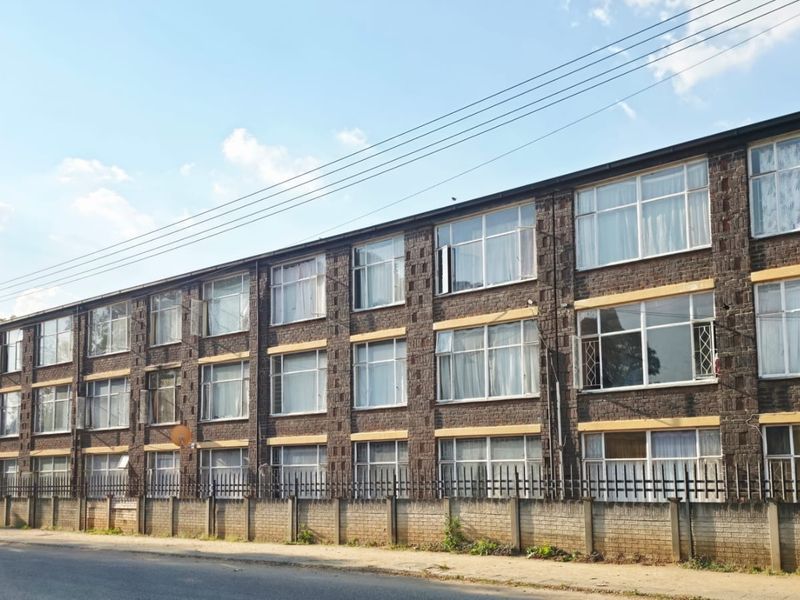 A multi-story residential apartment building featuring a classic brick facade, large metal-framed windows, and a secure perimeter boundary wall.