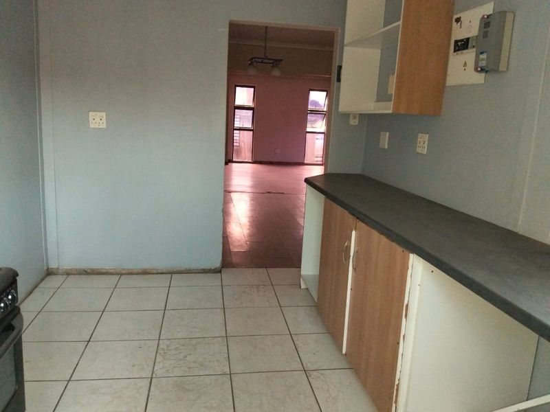 A functional kitchen space featuring tiled flooring, laminate cabinetry, and a dark countertop, leading into an adjacent living area.