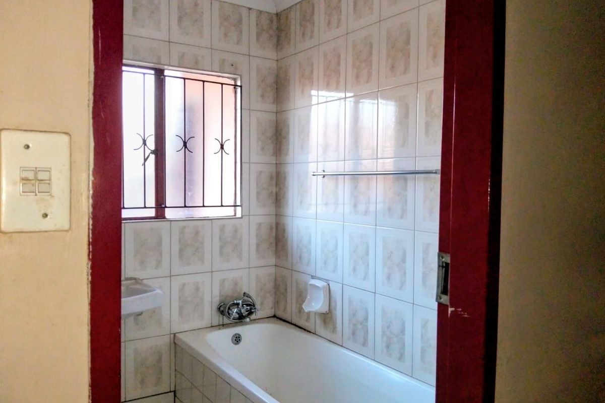 A dated bathroom featuring a standard bathtub, wall-mounted basin, and tiled walls with security bars on the window.