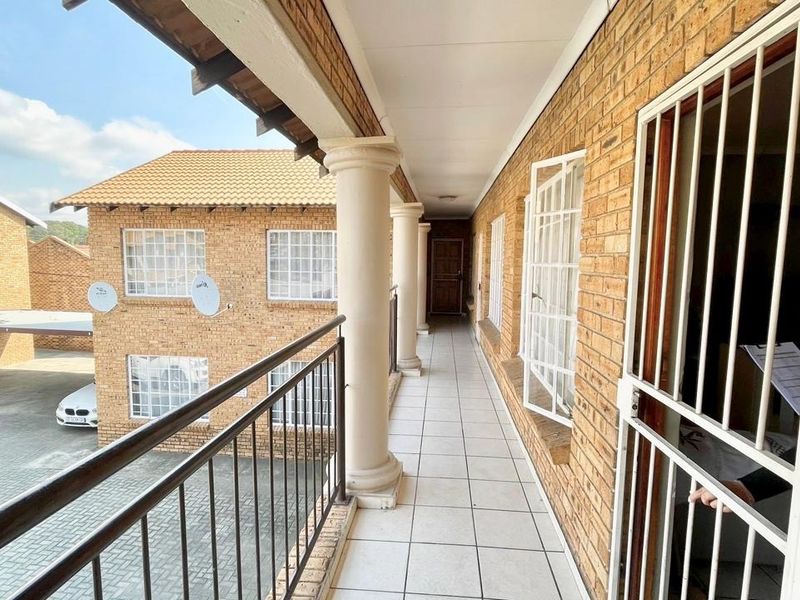 A covered exterior balcony featuring brick walls, tiled flooring, and metal security fixtures overlooking the apartment complex courtyard.