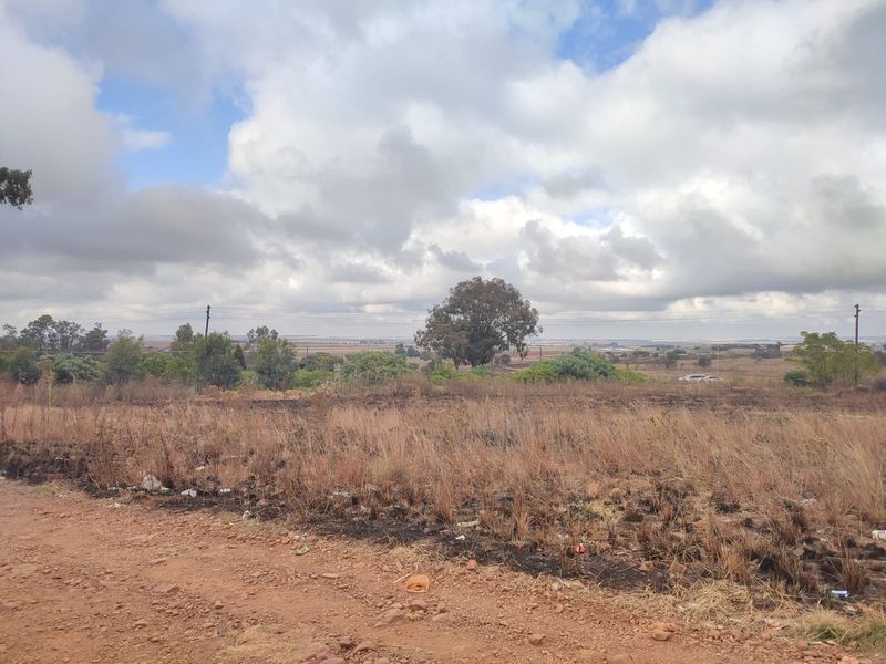 A view of a vacant residential plot featuring dry, scorched vegetation, dirt road access, and distant landscape views under a cloudy sky.