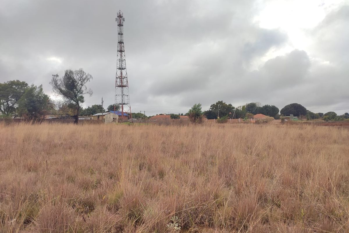 A view of a vacant residential plot featuring tall dry grass and a telecommunications tower in the background.