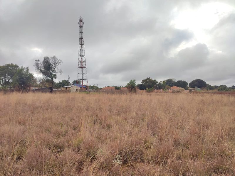A view of a vacant residential plot featuring tall dry grass and a telecommunications tower in the background.