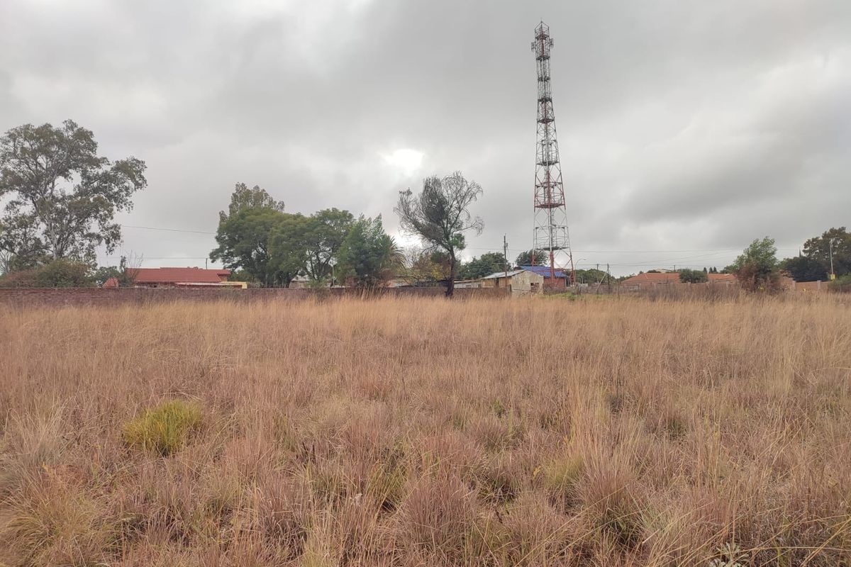 A vacant residential plot featuring tall dry grass, enclosed by a perimeter wall, with a telecommunications tower visible in the background.