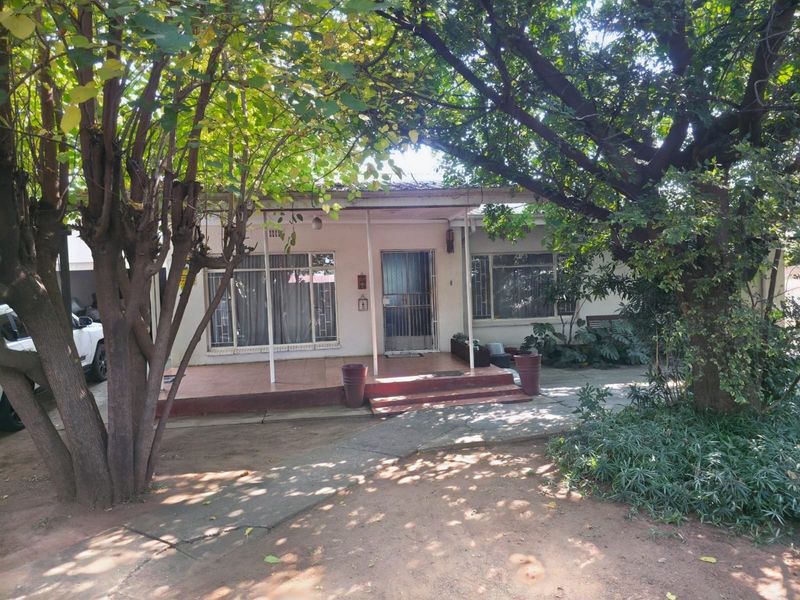 Front elevation of a single-story house featuring a covered patio, security gate, and mature trees in the front garden area.