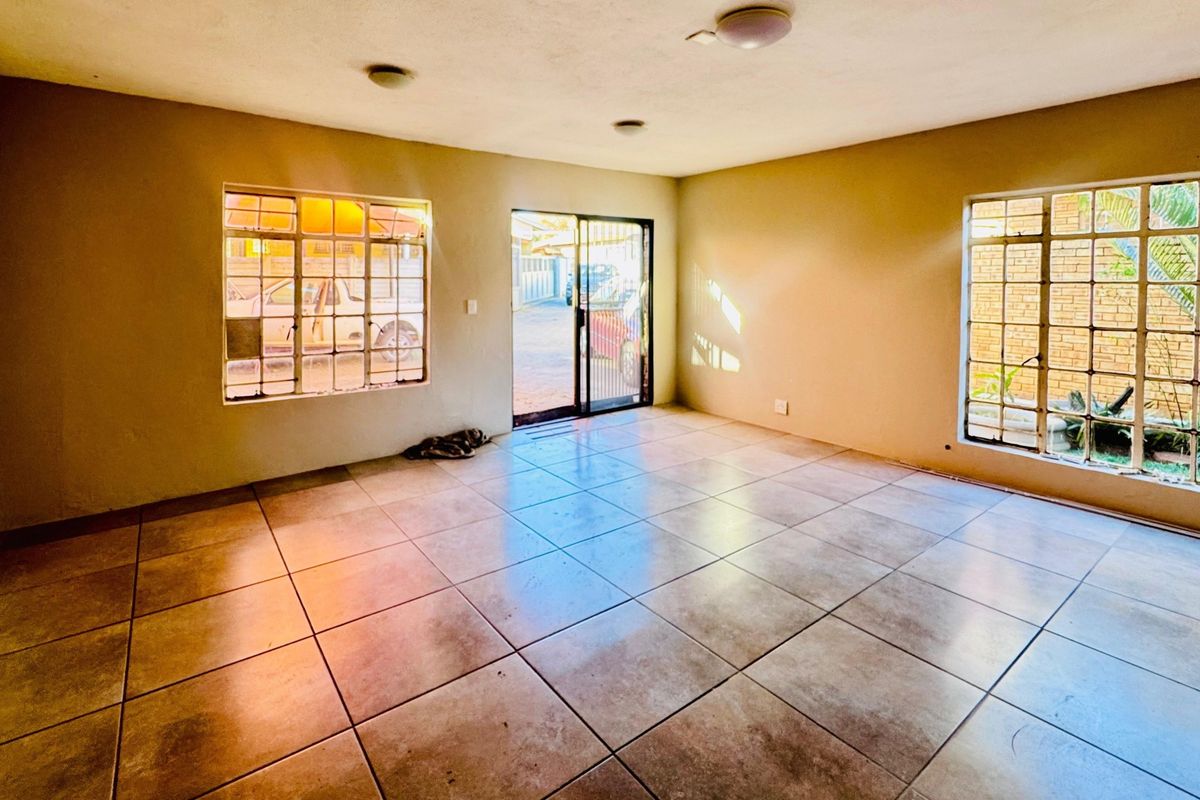 An empty, sunlit living room featuring large tiled floors, steel-framed windows, and a sliding glass door providing access to the exterior.