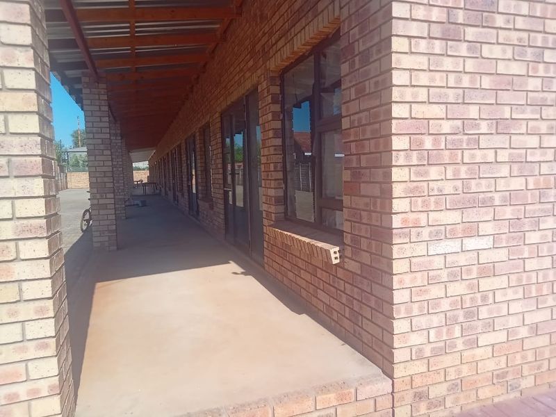 A covered outdoor walkway or patio area featuring exposed brick walls, concrete flooring, and aluminium-framed windows along the building exterior.