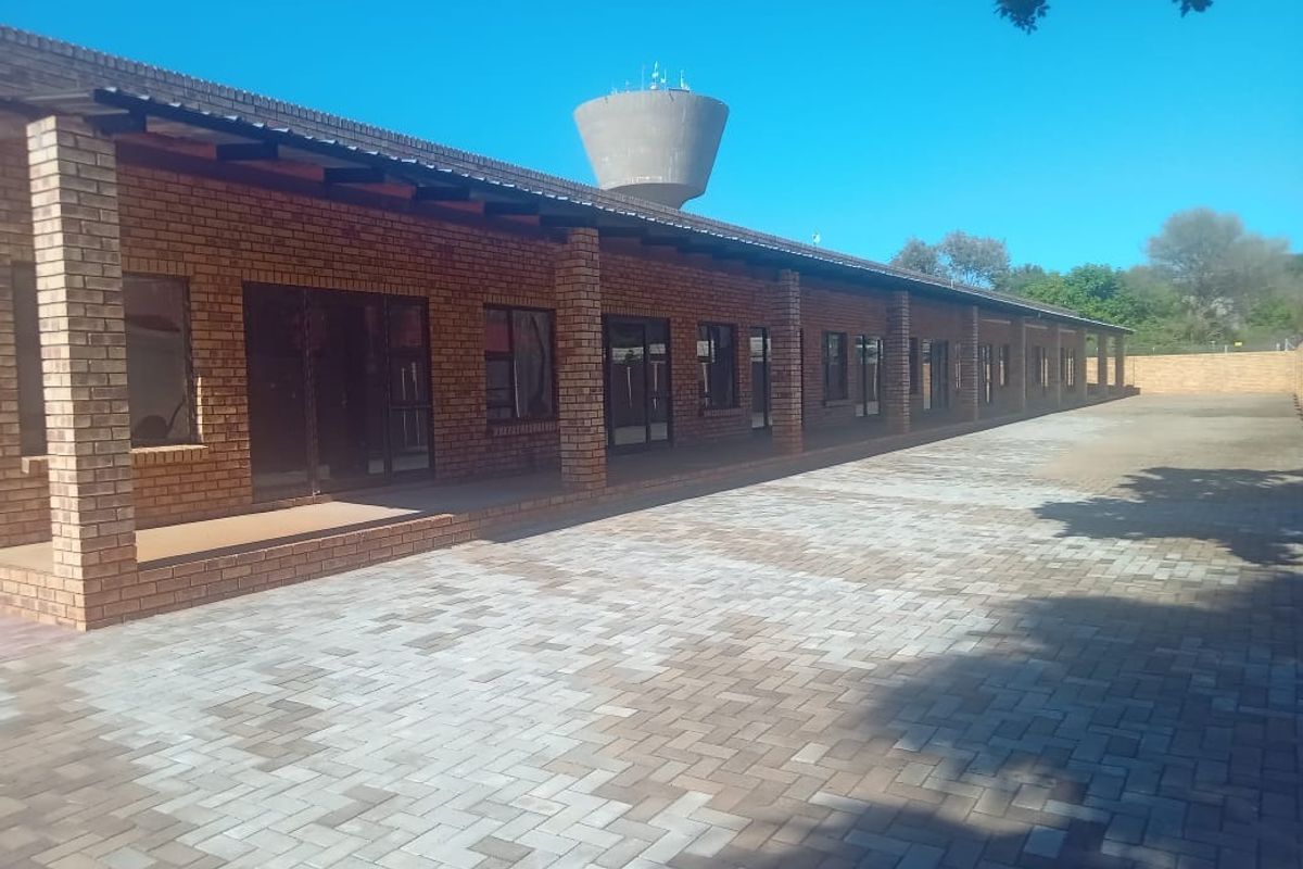 A spacious exterior rear courtyard featuring a long face brick building with a covered walkway, paved flooring, and multiple sliding glass door access points.