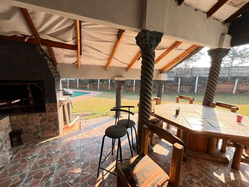 Covered outdoor patio featuring a built-in braai, decorative spiral columns, and tiled flooring, overlooking the garden and swimming pool area.
