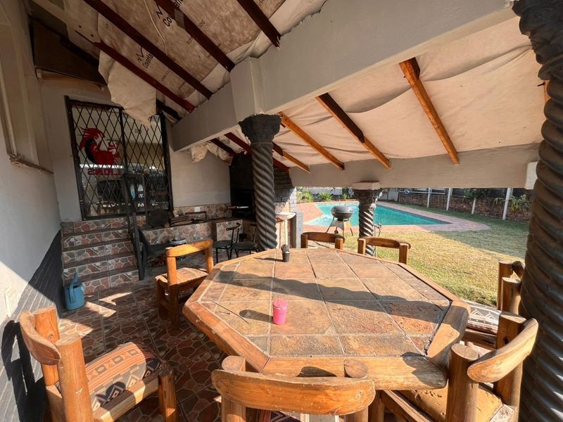 Covered outdoor patio area featuring a built-in braai, decorative columns, and tiled flooring, overlooking the swimming pool and garden.