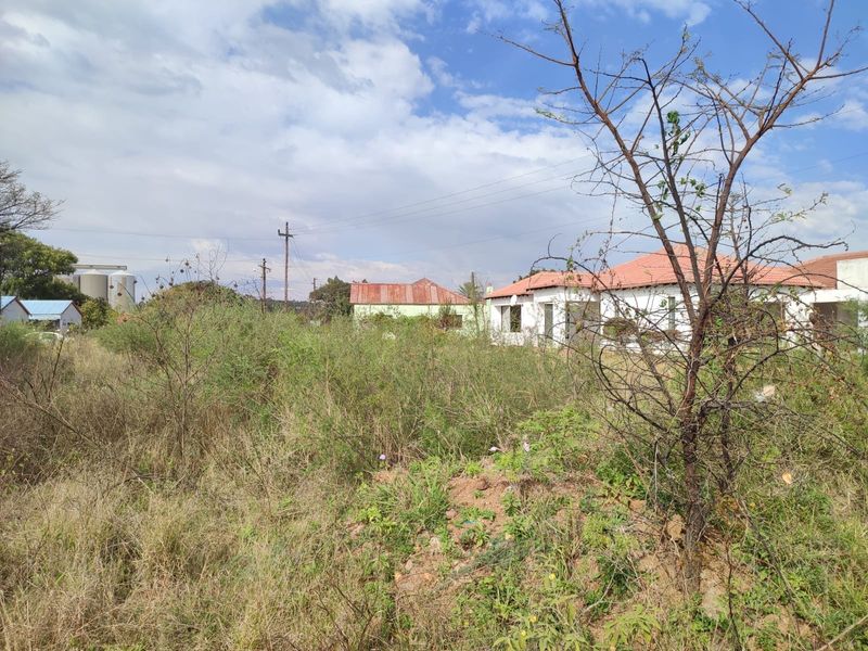 A view of a vacant residential plot featuring overgrown vegetation and existing structures in the background under a cloudy sky.