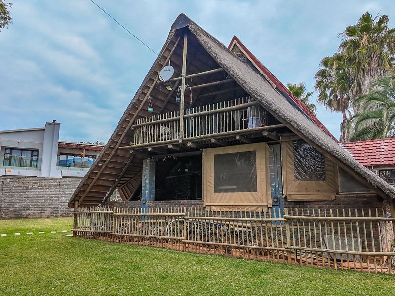 Rear view of an A-frame house featuring a thatched roof, timber balcony, and enclosed patio area set within a private garden.