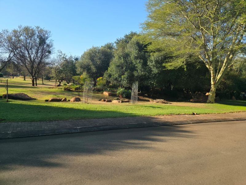 A well-maintained residential garden area featuring mature trees, natural rock formations, and a manicured lawn adjacent to a paved street.