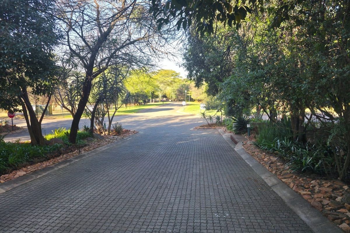 A view of a paved residential driveway leading towards a street intersection, surrounded by mature trees and established garden landscaping.